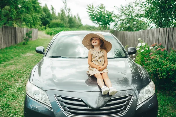 Carino bambina in cappello seduto sul cofano dell'auto, guardando ai lati . — Foto Stock