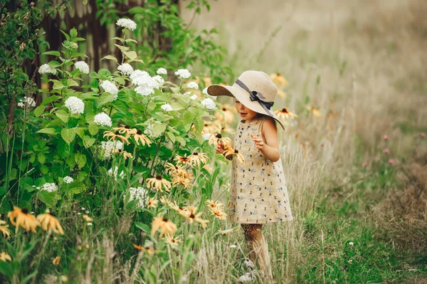 Linda chica en sombrero de paja en su cabeza oliendo flores en un jardín de verano . — Foto de Stock