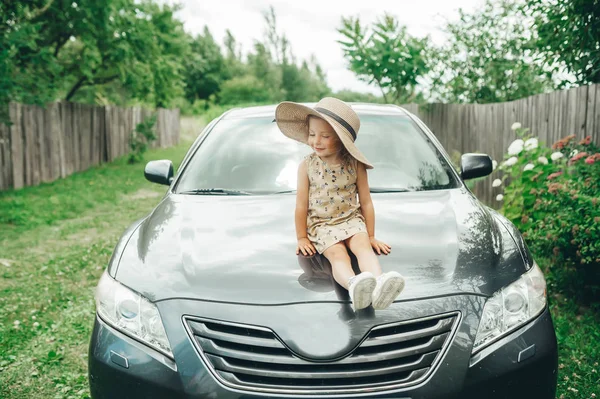 Carino bambina in cappello di paglia seduta sul cofano dell'auto nel villaggio — Foto Stock