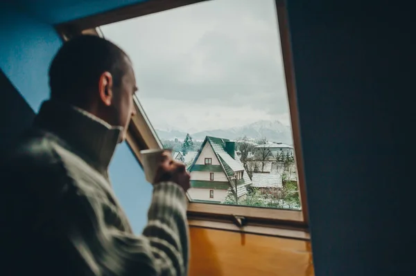 El hombre en jersey blanco con taza de café está de pie frente a la ventana y se ve hermosa ciudad —  Fotos de Stock