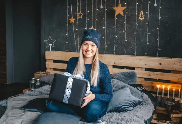 Блондинка в синей кепке, в голубых джинсах, с подарочной коробкой в руках, сидит на кровати в спальне . — стоковое фото