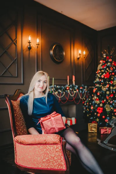 Портрет блондинки с подарком сидя в красном кресле рядом с праздничной елкой в коротком синем платье . — стоковое фото