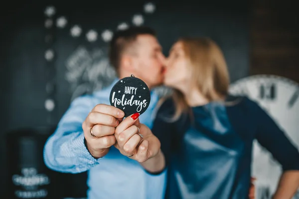 Küssendes Paar mit einem hölzernen runden Schild "Frohe Feiertage". Paar hält ein hölzernes rundes Schild "Frohe Feiertage". — Stockfoto