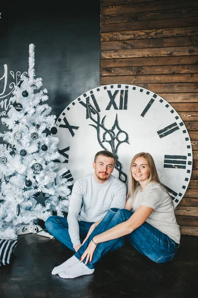 Pareja joven se sienta en el suelo cerca de abeto de Navidad en la habitación contra el fondo del gran reloj de pared — Foto de Stock