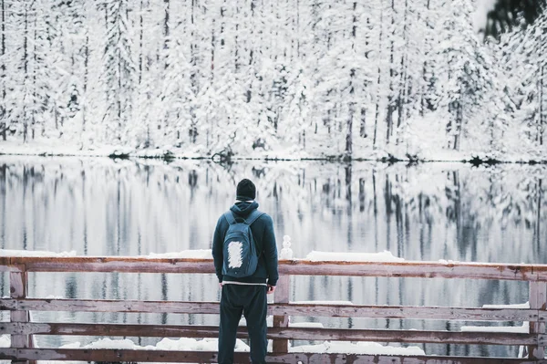 Un hombre viajero se queda en el lago de la montaña de nieve. El muñeco de nieve está cerca. Vista trasera . —  Fotos de Stock