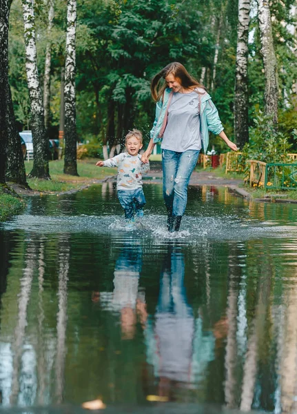 Feliz madre y su pequeño hijo en botas está corriendo a través de un charco después de la lluvia . —  Fotos de Stock