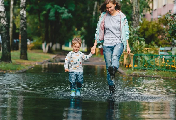 街の通りで雨が降った後、大きな水たまりの上を歩くレインブーツを着た彼女の小さな息子と幸せな母親. — ストック写真