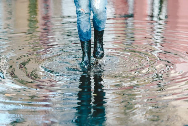 Obuwie damskie z czarnymi gumowymi butami i niebieskimi jeansami stojąc w kałuży wody po deszczu na ulicy miejskiej. Widok z przodu — Zdjęcie stockowe