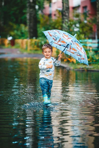 Menino feliz com guarda-chuva colorido brincando na poça em botas de borracha. Um menino se divertir depois de uma chuva . — Fotografia de Stock