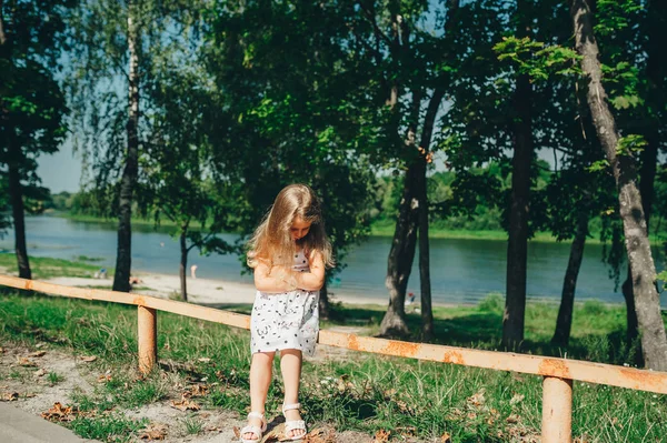 Mała dziewczynka w parku w pobliżu jeziora, przygnębiony siedzi na ławce. — Zdjęcie stockowe