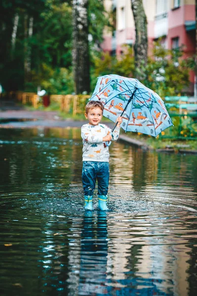 Menino adorável segurando guarda-chuva colorido em pé em uma poça no dia quente de verão . — Fotografia de Stock