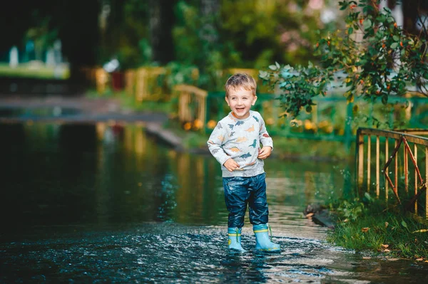 Dziecko po deszczu. Cute szczęśliwy chłopiec dziecko gra w kałuży. Dziecko w czarnych gumowych butach. — Zdjęcie stockowe