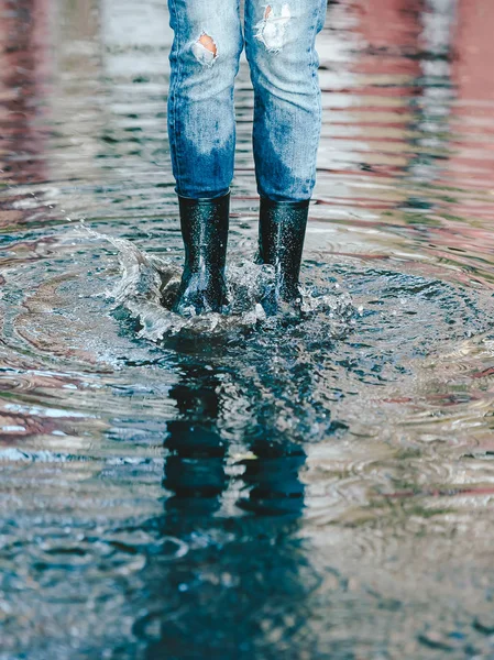 Pernas de mulher em jeans shabby com botas de borracha preta de pé em uma poça de água . — Fotografia de Stock