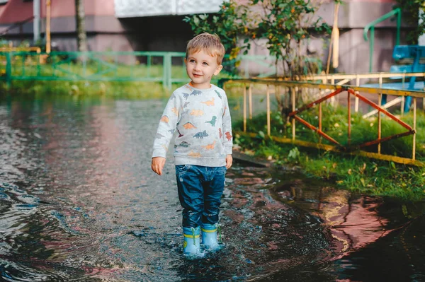 Mały chłopak po deszczu ubrany w gumowe buty stoją w wielkim kałuży. — Zdjęcie stockowe