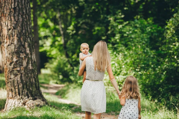 Hermosa madre de pelo largo en un vestido de verano en el bosque soleado pasea con sus hermosas hijas. Vista trasera . —  Fotos de Stock