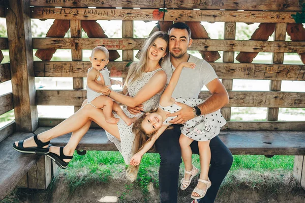 Retrato de una madre y un padre sonrientes sentados en un banco afuera con sus hijas jóvenes en gazebo en el parque . — Foto de Stock