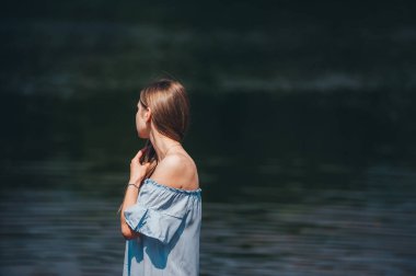 Beautiful sensual woman with long hair in blue dress posing on the lake shore on summer day