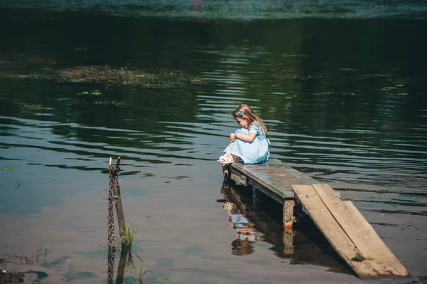 Zamyšlená holčička v modrých šatech sedí na malém dřevěném molu na pozadí jezera — Stock fotografie