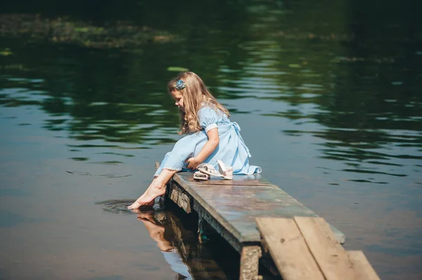 Una linda niña en vestido azul está sentada en un pequeño muelle de madera . — Foto de Stock