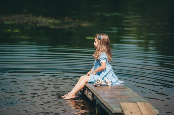 Dziewczynka w niebieskiej sukni siedzi na drewnianym molo w pobliżu wody. Kid sam na rzece. Widok z boku — Zdjęcie stockowe
