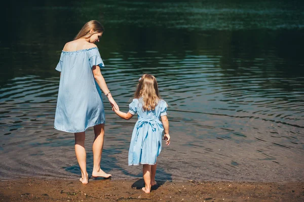 La joven y su hija descalzas caminan en el agua sobre un río. Vista trasera —  Fotos de Stock