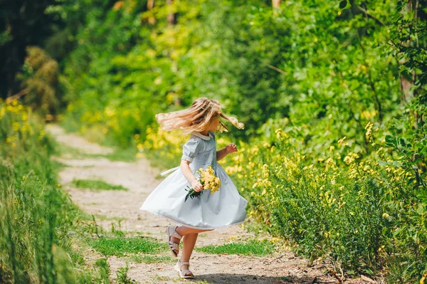 Niña en vestido azul con flores silvestres amarillas correr por un camino en el bosque — Foto de Stock