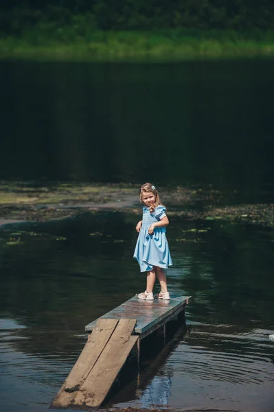 Sonriente niña en vestido azul mira a la cámara en el muelle cerca del lago . —  Fotos de Stock