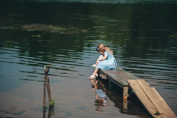 Holčička v krásných šatech sedí na malém dřevěném molu na pozadí jezera — Stock fotografie