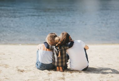 Nehir kıyısında kum üzerinde oturan üç kişilik bir aile. Sevimli oğlu anne ve babasını kucaklayan. Arkadan görüntüle.