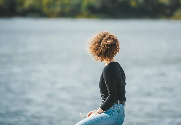 Mujer joven con el pelo largo rizado y jeans sentados con la espalda mirando el paisaje del agua . — Foto de Stock