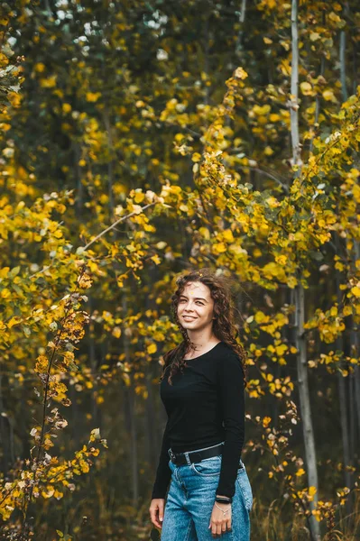 Красива стильна дівчина в чорній сорочці і в блакитних джинсах стоїть і позує в осінньому парку . — стокове фото