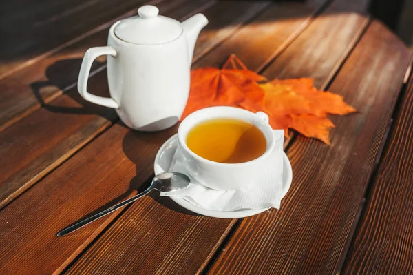 White tea cup with black tea, white teapot on wooden table and autumn maple leaves.