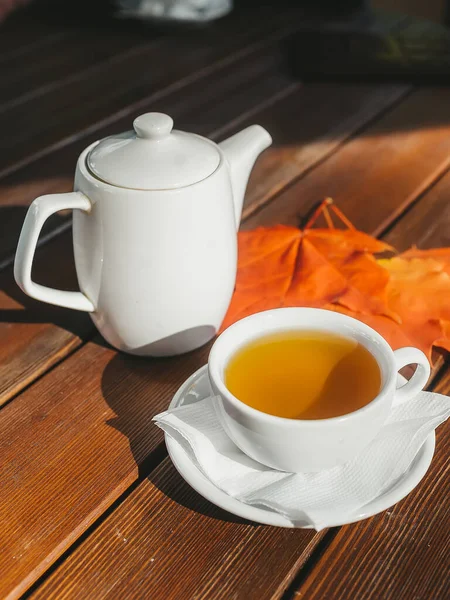 White tea cup with black tea, white teapot on wooden table and autumn maple leaves.