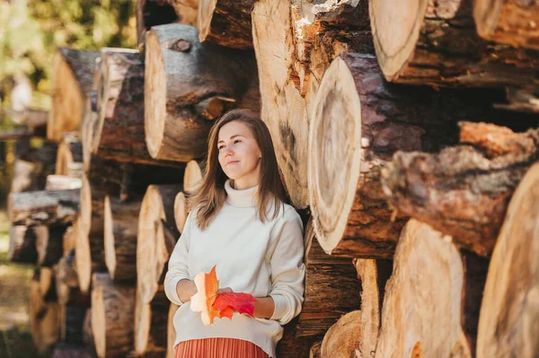 Dreaming kobieta w białym swetrze z liści klonu w rękach stoi na tle drewna na opał i kłody. — Zdjęcie stockowe