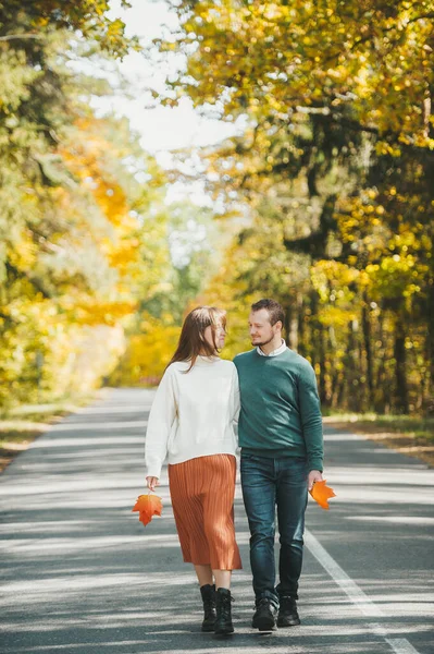 Amar casal feliz no outono no parque andando na estrada de asfalto e segurando folhas de bordo de outono em mãos — Fotografia de Stock