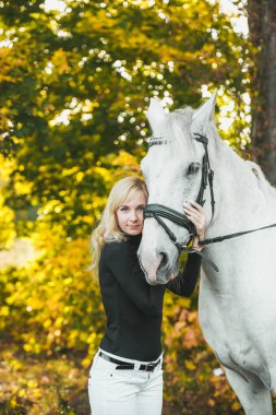 Genç ve çekici sarışın bir kadın sonbahar ormanında beyaz bir ata sarılıyor..