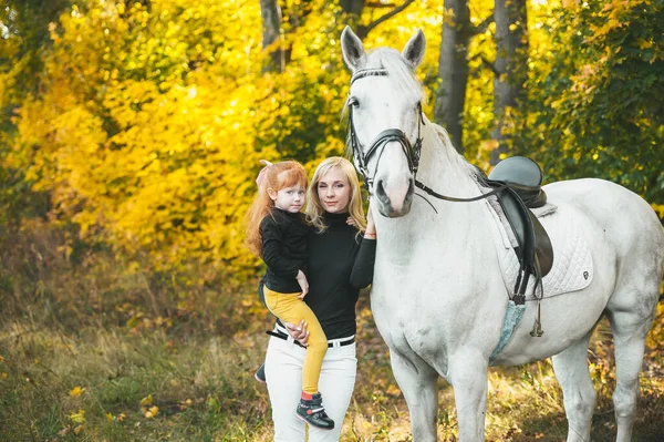 En ung mor med sin rödhåriga lilla dotter och vita en häst poserar i höstskogen. — Stockfoto