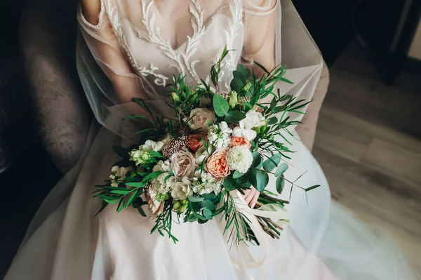 Die Braut sitzt auf dem Sofa und hält einen Brautstrauß aus Rosen in einem Brautkleid. Nahaufnahme — Stockfoto