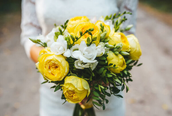 A caucasian woman is standing outdoors, holding a bridal bouquet of yellow roses wearing a wedding dress.