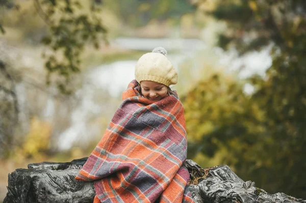 Pequeña niña sonriente envuelta en una manta está sentada en talón de madera cerca del lago en un parque de otoño — Foto de Stock