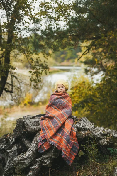 Mała dziewczynka owinięta w koc siedzi na drewnianym kawałku w pobliżu jeziora w jesiennym lesie — Zdjęcie stockowe