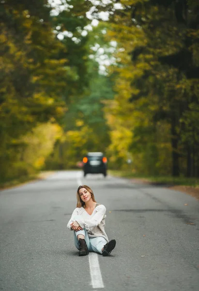 Szczęśliwa kobieta w dżinsach siedzi w centrum na asfaltowej drodze w jesiennym lesie — Zdjęcie stockowe