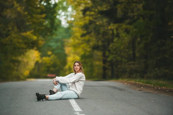 Szczęśliwa kobieta w dżinsach siedzi w centrum na asfaltowej drodze w jesiennym lesie — Zdjęcie stockowe