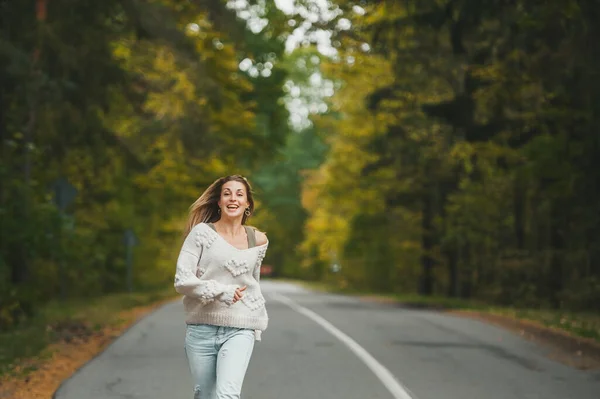 在秋天的森林里，穿着牛仔裤的快乐女人正在柏油路上奔跑 — 图库照片