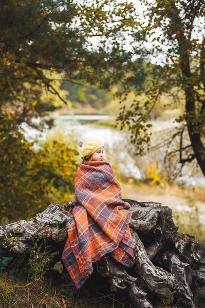 La niña seria en manta caliente contra el fondo del hermoso lago del bosque al aire libre . — Foto de Stock