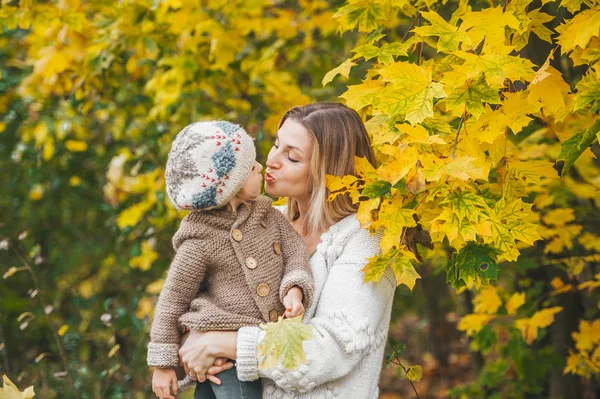 Mother kissing and hugging her beautiful little daughter while standing near maple tree in autumn park.