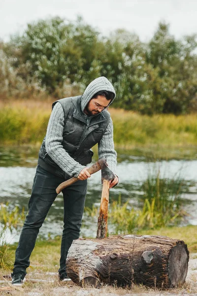 Человек в очках раскалывает дрова и рубит дрова топором на берегу реки . — стоковое фото