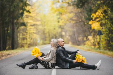 Sonbahar günü asfalt yolda sırt sırta oturan bir çift. Sarı akçaağaç yaprakları buketleriyle boş bir yolda genç bir çift..