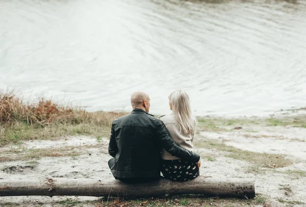 男人和一个女孩正靠着他们的背坐在湖边 这对浪漫的夫妇坐在湖边或河边的圆木上 — 图库照片