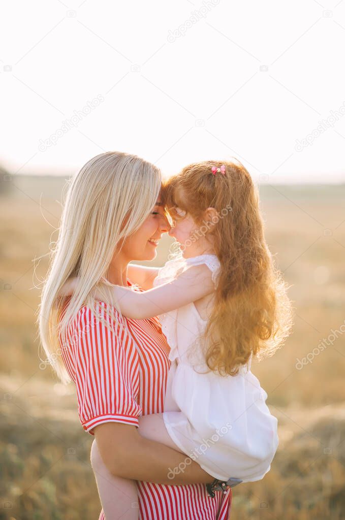 Emotional portrait of a happy and active young blonde mother holding her positive little red-heired daughter in her arms while standing in a field in the sunset light. Summer life